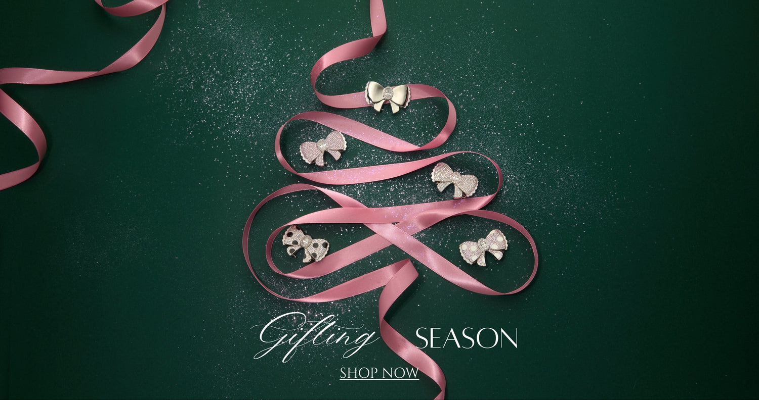 Pink ribbon shaped like a Christmas tree with jewelry on a green background, promoting the 'Gifting Season'. Charmante Bow Bag Charm