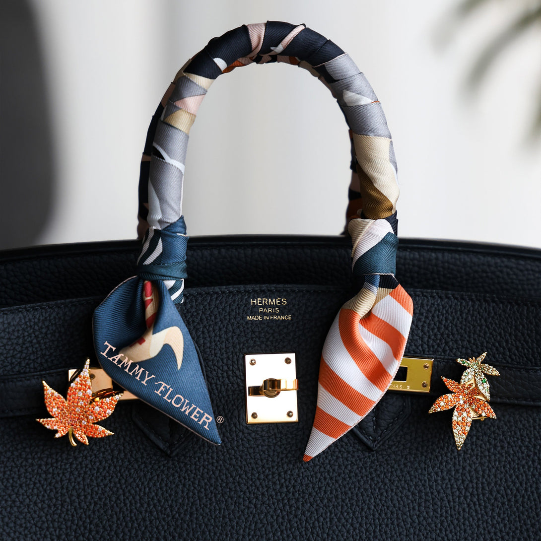 Black birkin 25 handbag with colorful scarf and decorative maple leaf pins on a light background