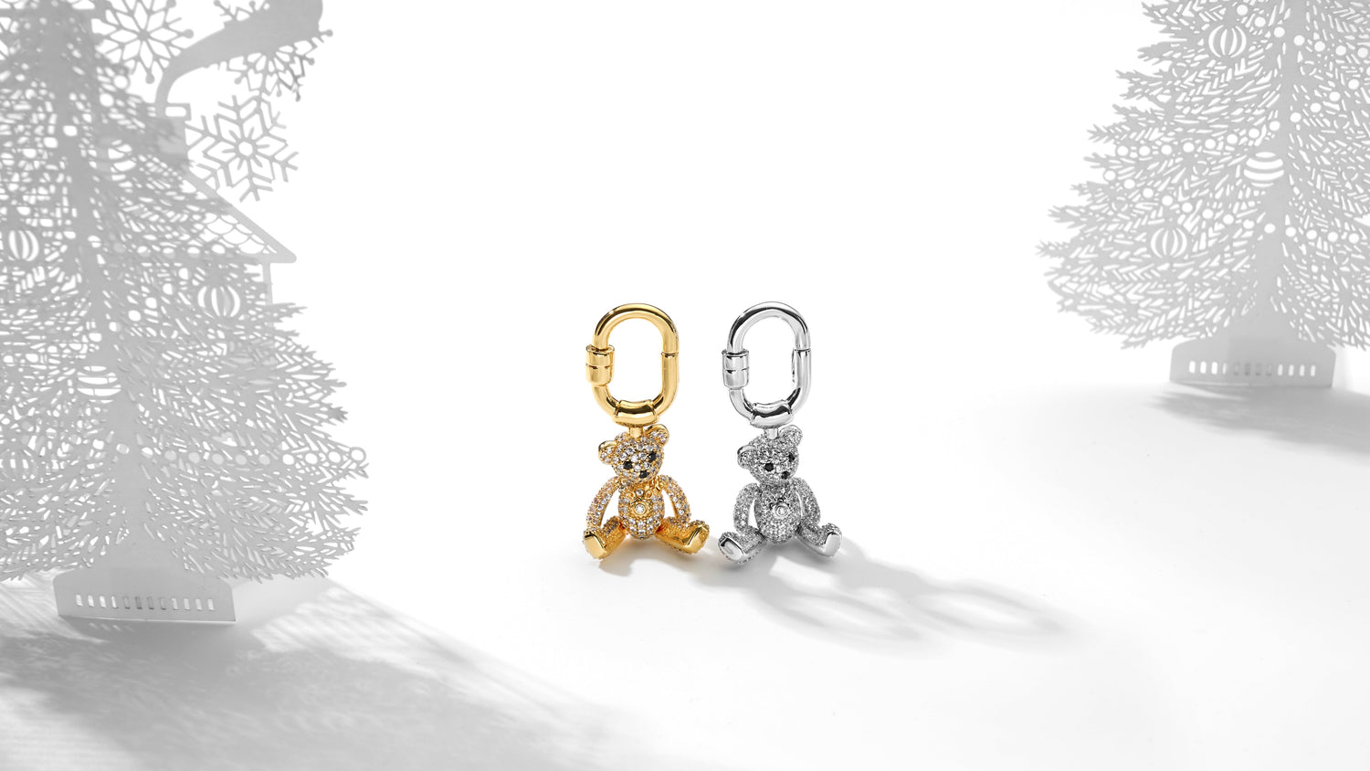 Two teddy bear-shaped jewelry bag charms, one gold and one silver, on a white surface with decorative trees in the background.